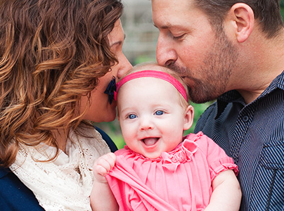 Two adults, facing each other, kissing the head of a smiling baby held between them in their arms.