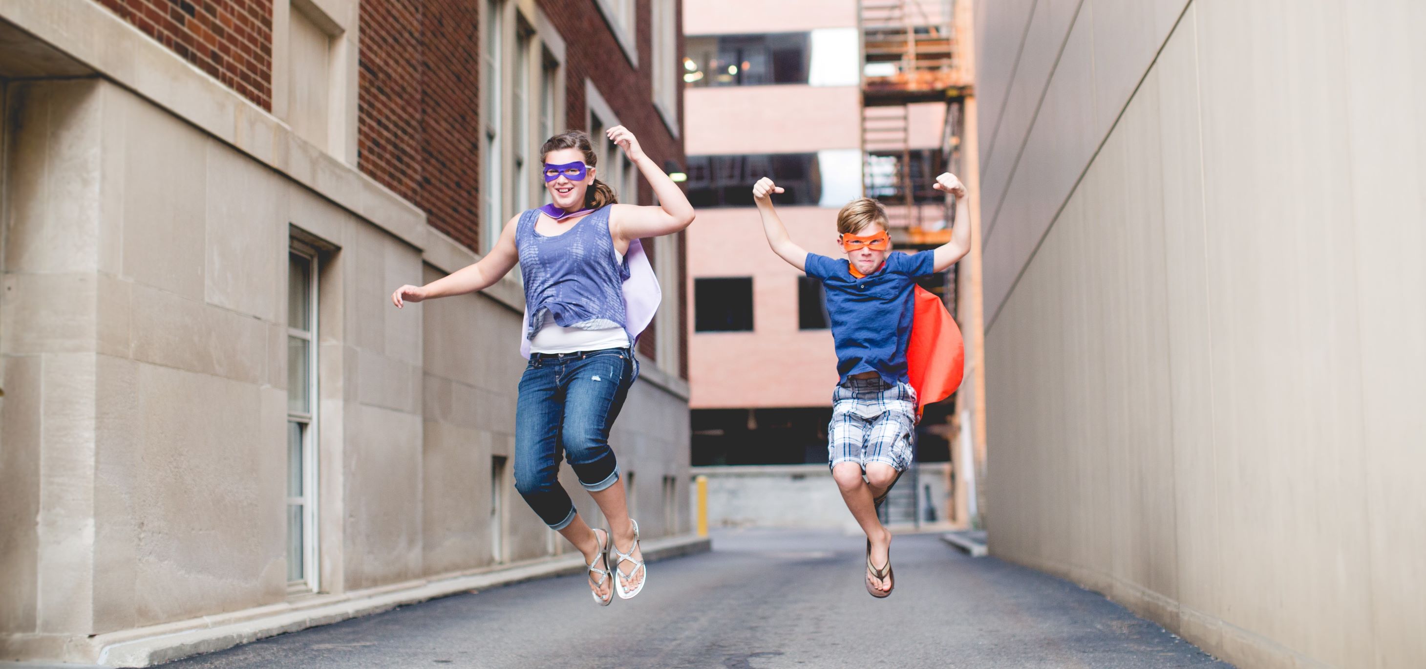 Older sister and younger brother leaping into the air as superheroes with masks and capes